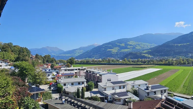 Panoramablick über eine Wohnsiedlung mit Feldern und majestätischen Bergen im Hintergrund unter blauem Himmel.