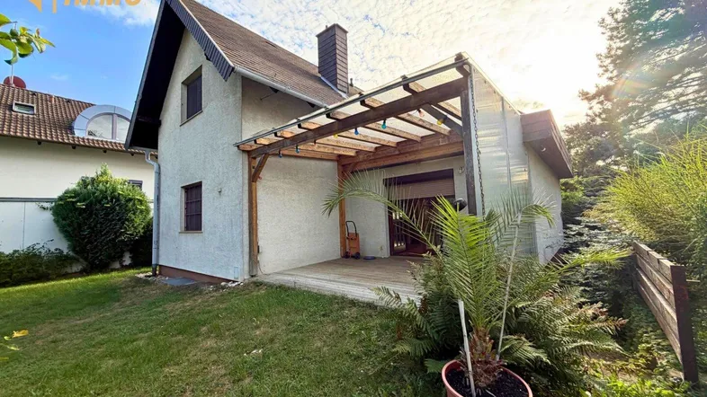 Hinteransicht des Hauses mit überdachter Holzterrasse und grünem Gartenbereich unter strahlendem Himmel.