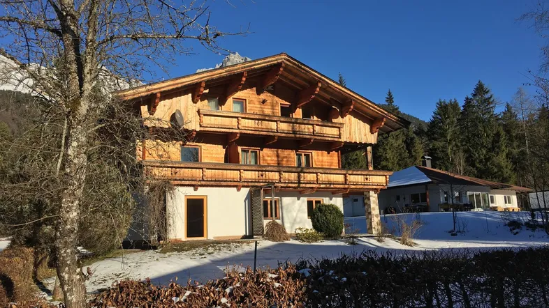 Traditionelles Holzchalet mit mehreren Balkonen und verschneiter Umgebung vor Bergkulisse an einem sonnigen Tag.