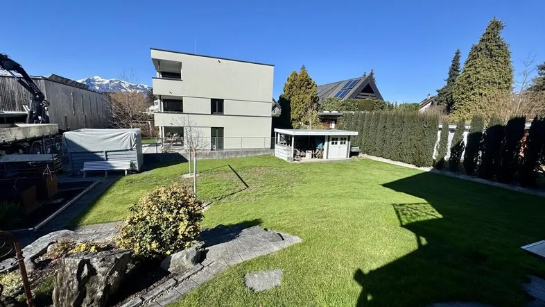 Gepflegter Garten mit Rasenfläche, Gartenhaus und modernem Gebäude im Hintergrund mit Bergblick.