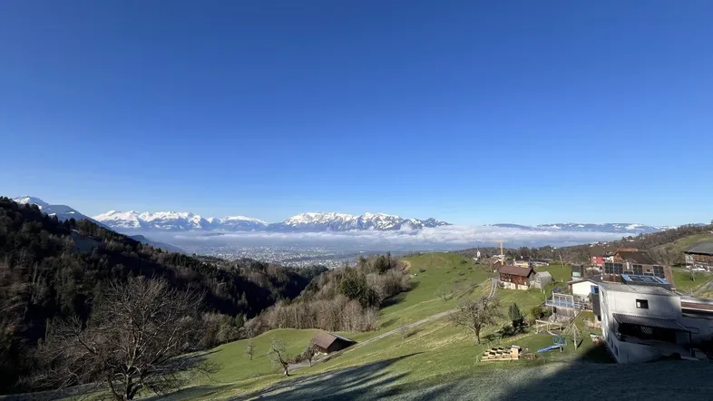 Panoramablick über ein grünes Tal mit einer Stadt in der Ferne und schneebedeckten Bergen unter blauem Himmel.