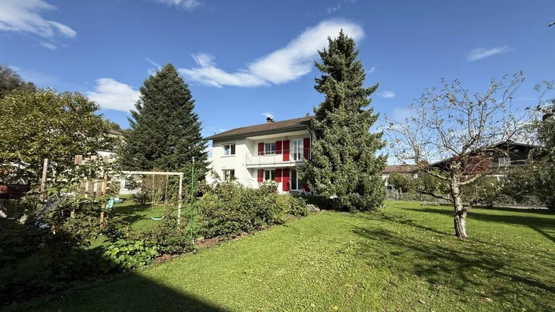 Großzügiger Gartenbereich mit Rasenfläche, Bäumen und einem Schaukelgerüst unter blauem Himmel.