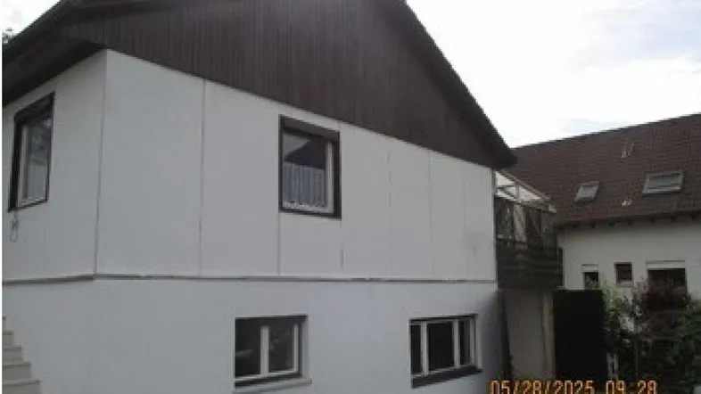 Ansicht des Hauses von außen mit weißer Fassade, braunem Dach und Fenstern mit Rollläden.