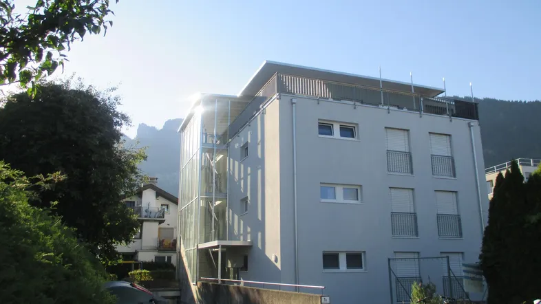 Modernes Mehrfamilienhaus mit heller Fassade, Balkonen und einem gläsernen Treppenhaus vor Bergkulisse.