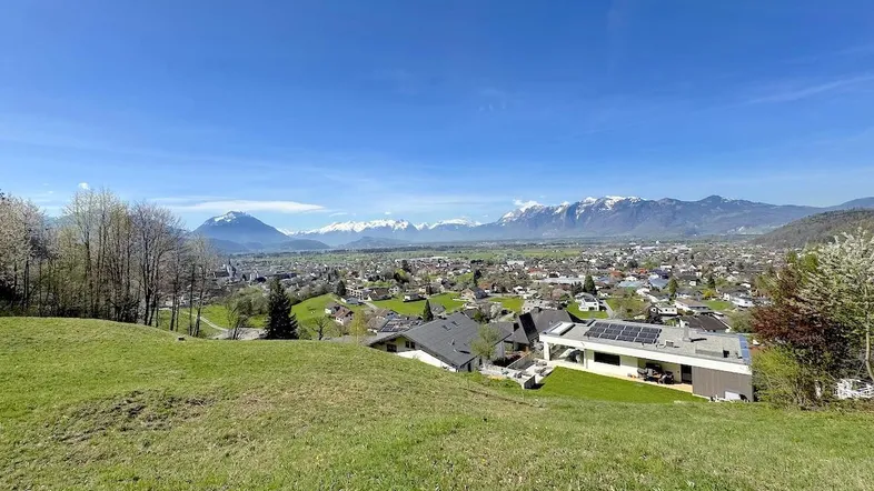 Panoramablick über eine Stadt im Tal mit majestätischen Bergen im Hintergrund unter blauem Himmel.