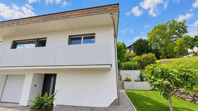 Moderne Hausfassade mit Garage, Balkon und gepflegtem Gartenbereich am Hang mit grüner Aussicht.
