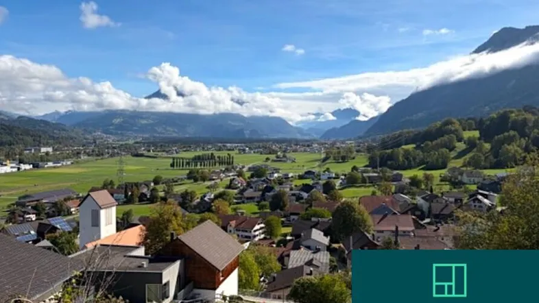 Panoramablick auf ein Dorf in einem Tal mit Bergen im Hintergrund und weitem Himmel.