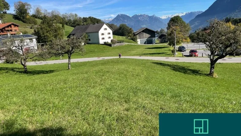 Grünes Grundstück mit Blick auf die umliegende Berglandschaft und Wohnhäuser unter blauem Himmel.