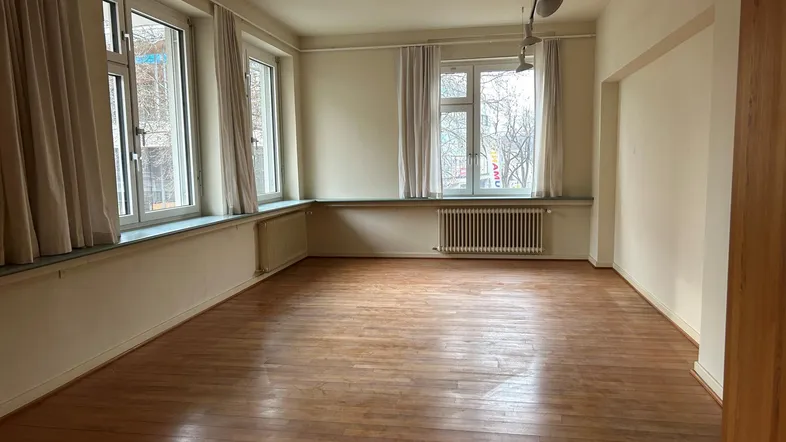 Geräumiger, leerer Raum mit Holzboden und großen Fenstern, die viel Tageslicht hereinlassen.