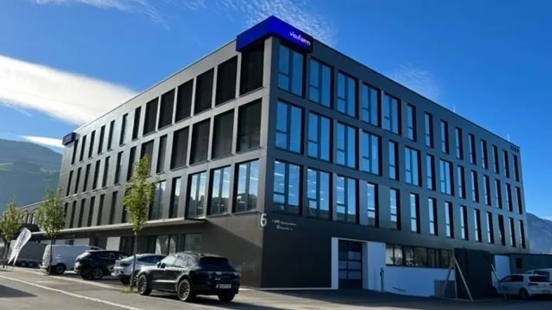 Modernes Bürogebäude mit dunkler Fassade, großen Fenstern und blauer Beschriftung unter klarem Himmel.