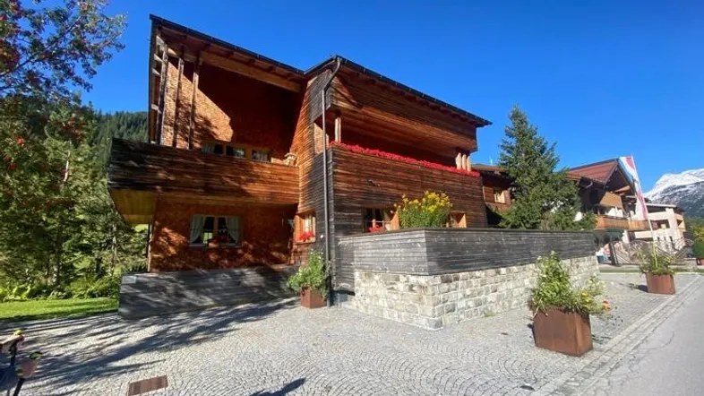 Modernes Chalet mit Holzfassade, gepflastertem Vorplatz und Balkonen mit Blumenkästen vor alpiner Kulisse.