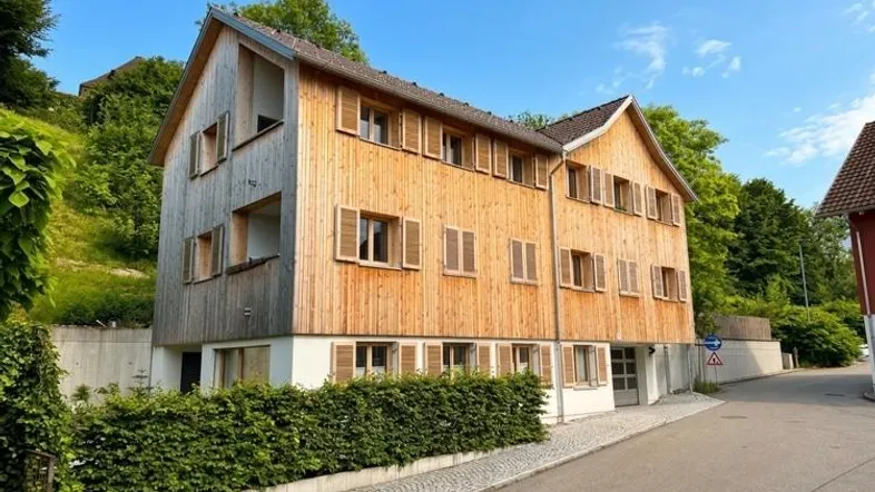 Modernes Mehrfamilienhaus mit Holzfassade und traditionellen Fensterläden, eingebettet in eine grüne Hanglage.