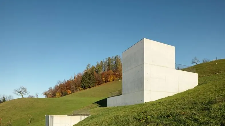 Modernes Gebäude aus Sichtbeton in einer grünen Hügellandschaft unter blauem Himmel.