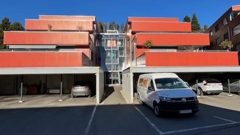 Modernes Gebäude mit leuchtend orangefarbenen Balkonen, Tiefgaragenzufahrt und zentralem Glasaufzug.