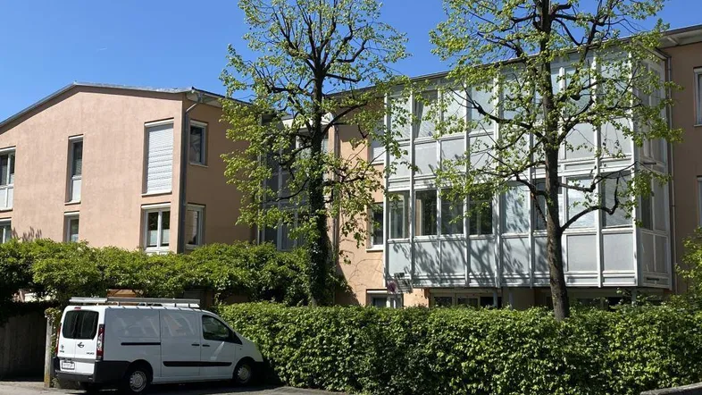 Mehrfamilienhaus mit verglasten Balkonen und gepflegter Hecke im Vordergrund, davor ein Parkplatz.