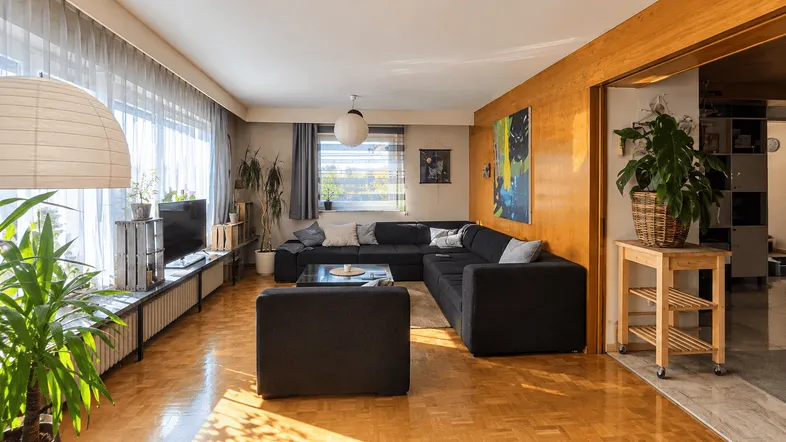 Helles Wohnzimmer mit dunklem Ecksofa, Parkettboden und Holzvertäfelung, umgeben von Pflanzen.