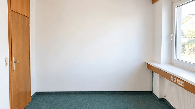 Heller Büroraum mit Holzdecke, weißen Wänden und grünem Teppichboden, ausgestattet mit einem Fenster.
