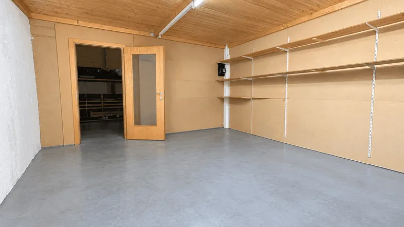 Geräumiger Lagerraum mit Holzdecke, robustem Betonboden und praktischen Regalsystemen an der Wand.