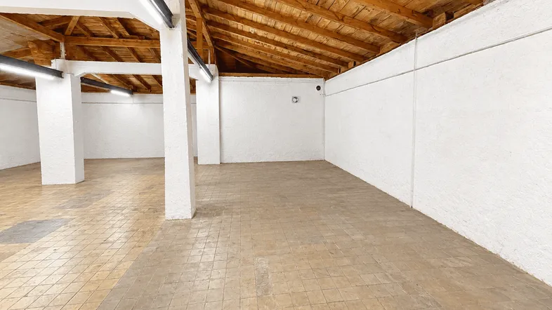 Geräumiger, leerer Gewerberaum mit sichtbaren Holzbalken und Fliesenboden unter einem schrägen Dach.