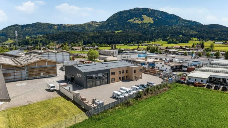 Luftaufnahme eines modernen Gewerbegebäudes mit umliegenden Grünflächen und Bergen im Hintergrund unter blauem Himmel.