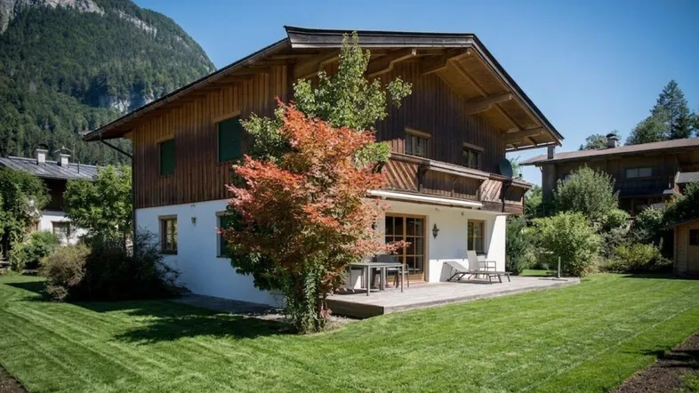Traditionelles Holzhaus mit weißer Fassade, großer Rasenfläche und einer Holzterrasse mit Bergblick.