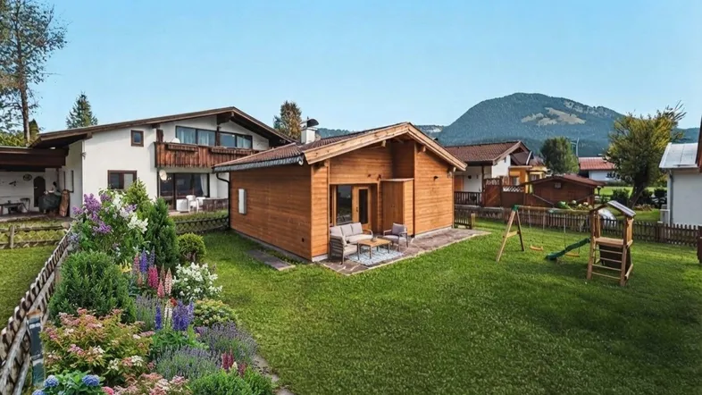 Gepflegter Gartenbereich mit Blumenbeeten, Rasenfläche, Spielplatz und einem kleinen Holzhaus mit Sitzgelegenheit.