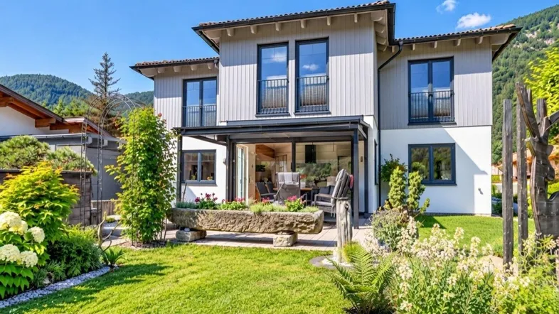Modernes Haus mit gepflegtem Garten, überdachter Terrasse und Bergblick unter blauem Himmel.