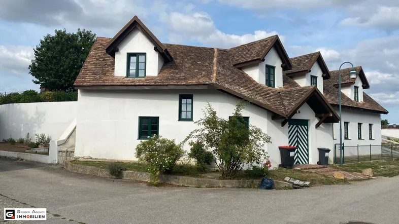 Weißes Landhaus mit traditionellem Ziegeldach und grünen Fensterläden, umgeben von Büschen.