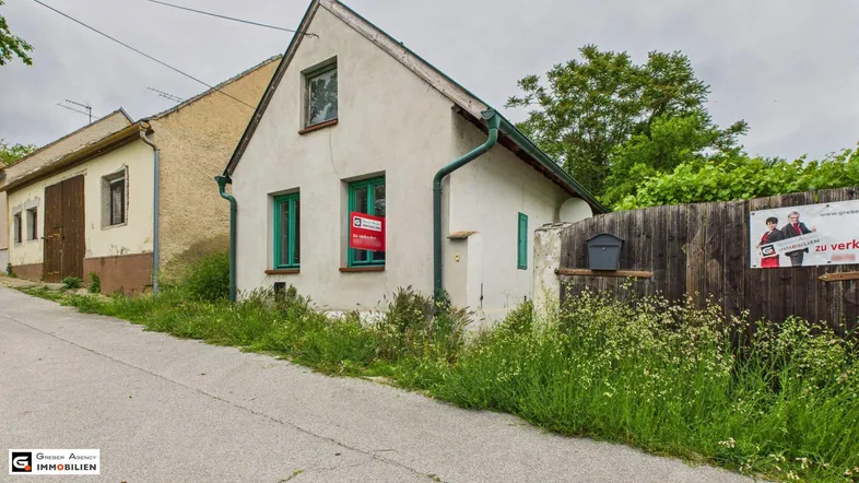 Altes, einfaches Haus mit weißer Fassade, grünen Fensterrahmen und einem "Zu verkaufen"-Schild.