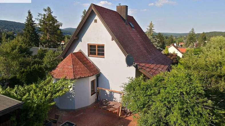 Freistehendes Einfamilienhaus mit rotem Ziegeldach und umgebender grüner Landschaft unter blauem Himmel.