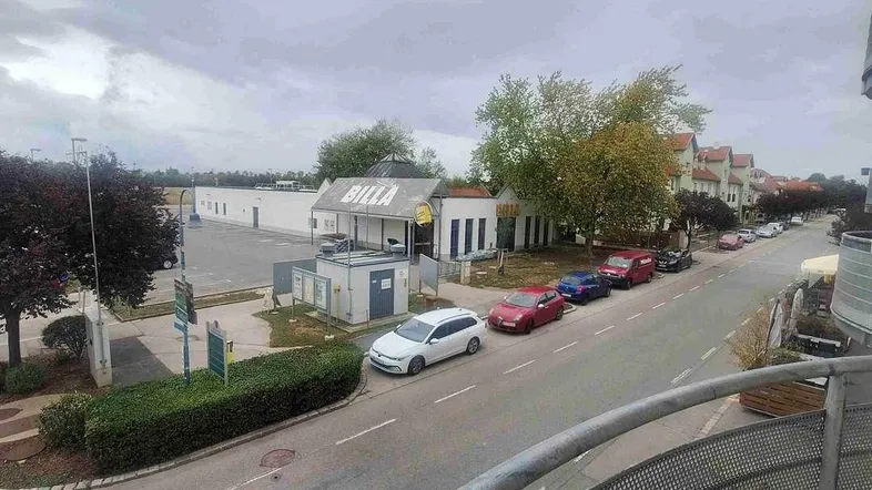 Blick vom Balkon auf die Straße mit parkenden Autos und einem Supermarkt in der Ferne unter bewölktem Himmel.