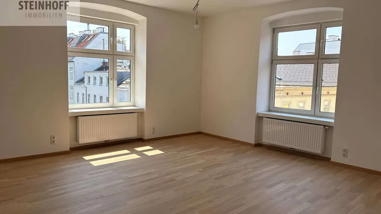 Helles, leeres Zimmer mit Parkettboden und zwei großen Fenstern, die viel Tageslicht hereinlassen.