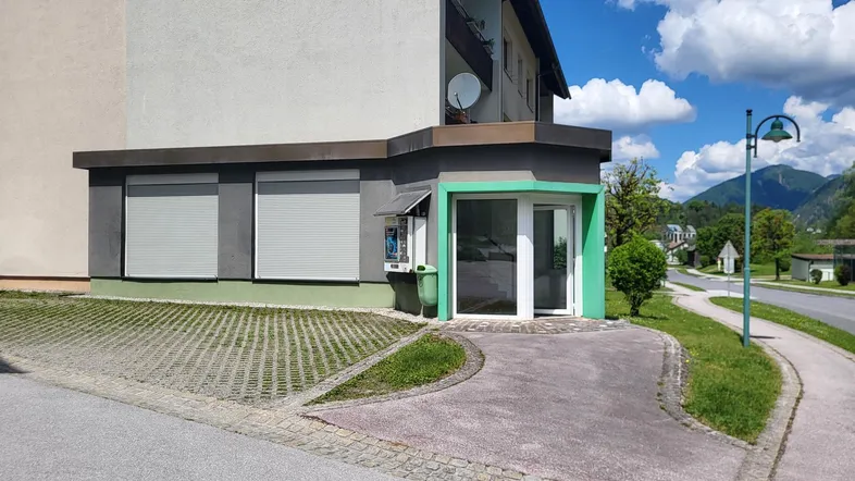 Gebäudeansicht mit Ladenfront, Rollläden und gepflastertem Vorplatz unter blauem Himmel.