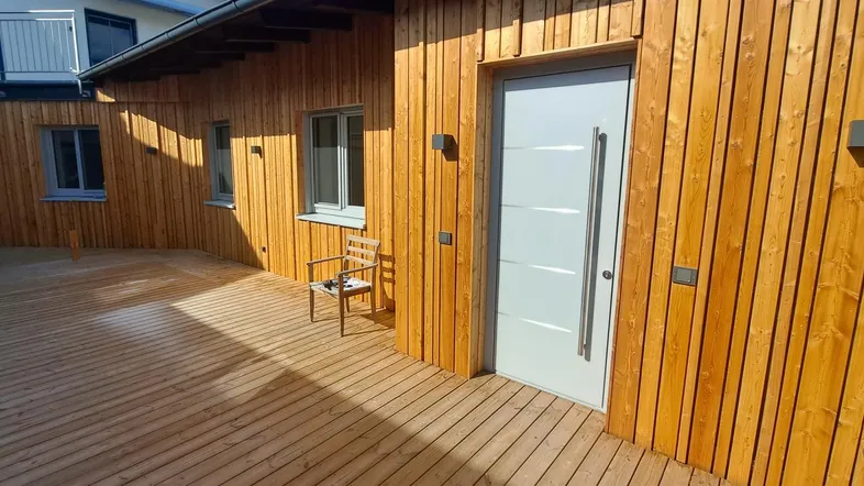Geräumige Holzterrasse mit moderner Eingangstür und Holzfassade des Gebäudes unter blauem Himmel.