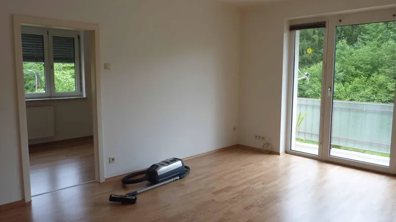 Heller Wohnraum mit Parkettboden, weiß gestrichenen Wänden und Zugang zum Balkon mit Grünblick.