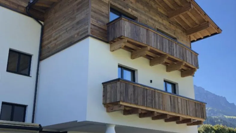 Modernes Gebäude im alpinen Stil mit Holzverkleidung und Balkonen, umgeben von grüner Landschaft.