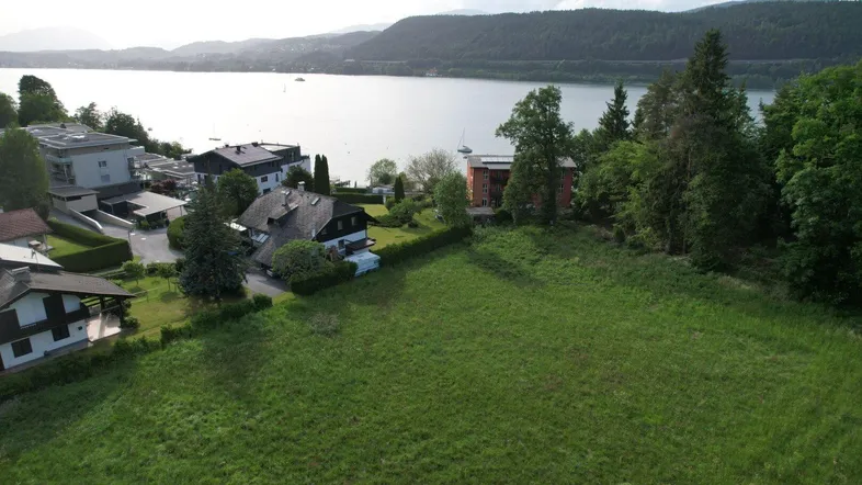 Luftaufnahme eines großen Grundstücks in Seenähe mit umliegenden Häusern und Bergen im Hintergrund.