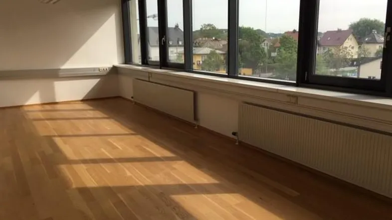 Heller Büroraum mit großen Fenstern, Holzboden und Blick auf die umliegende Bebauung.