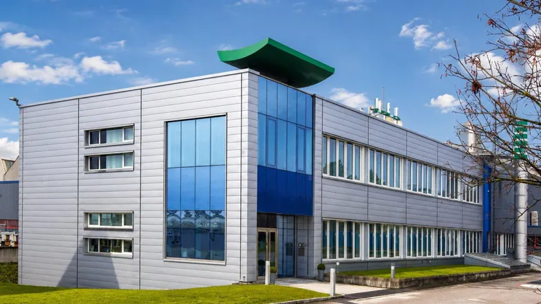 Modernes Bürogebäude mit grauer Fassade, großen blauen Glasflächen und einem markanten grünen Dachelement.