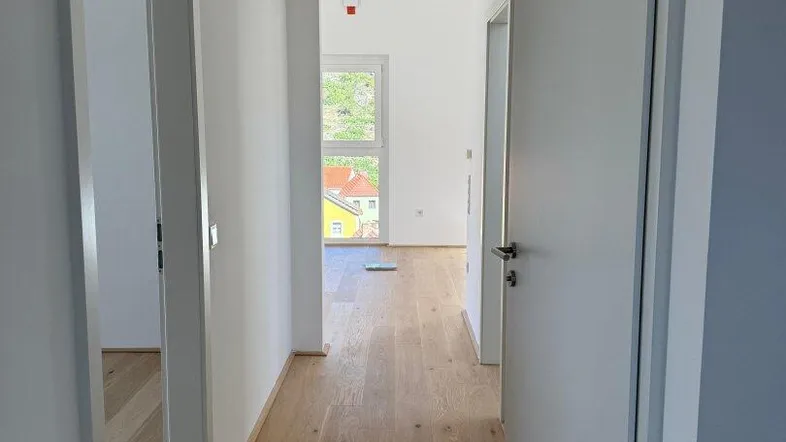 Heller Flur mit neuem Holzfußboden und Blick auf die grüne Umgebung durch das Fenster am Ende.