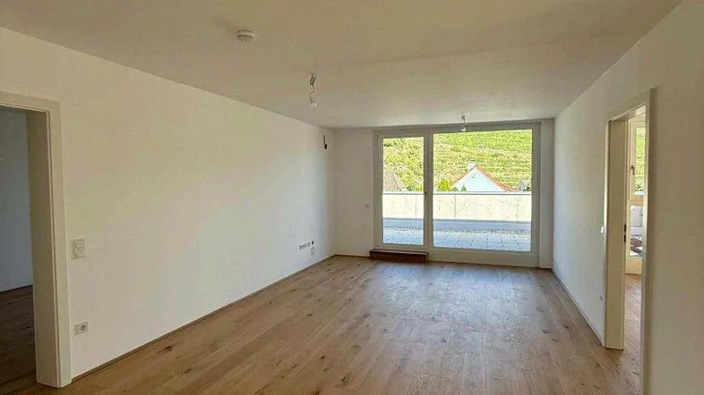 Heller, leerer Raum mit neuem Holzboden und großen Fenstern, die einen Blick ins Grüne bieten.
