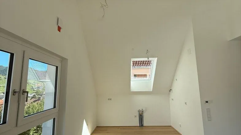 Heller Dachgeschossraum mit Holzbalken, großem Fenster und Dachfenster, bietet einen weiten Blick.