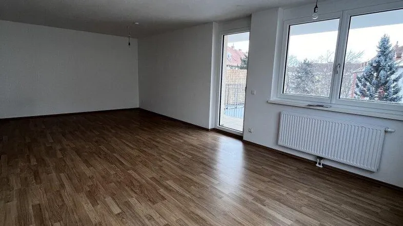 Geräumiges, leeres Wohnzimmer mit Laminatboden, großen Fenstern und Zugang zum Balkon.