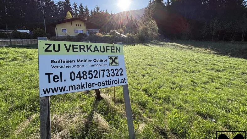 Verkaufsschild auf einer grünen Wiese mit einem Haus und Wald im Hintergrund bei Sonnenuntergang.