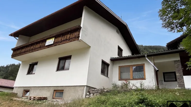 Traditionelles Haus mit Holzbalkon und weißer Fassade, umgeben von grüner Landschaft und Bergen.