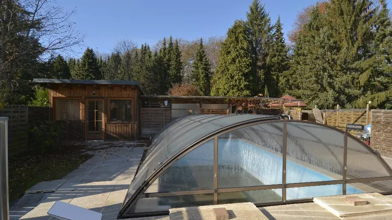 Poolbereich mit Überdachung und angrenzendem Holzhaus unter blauem Himmel.