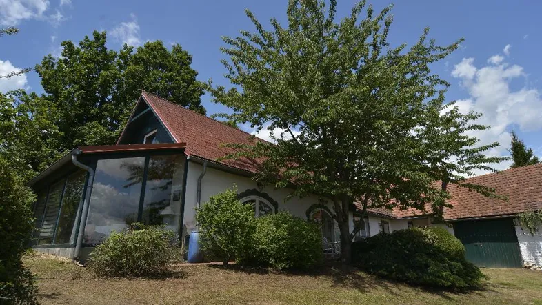 Hausfassade mit großem Wintergarten, umgeben von Bäumen und einem gepflegten Rasen unter blauem Himmel.