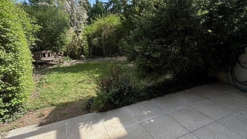 Geflieste Terrasse mit Pergola und Zugang zu einem grünen Gartenbereich mit Bäumen und Sträuchern.