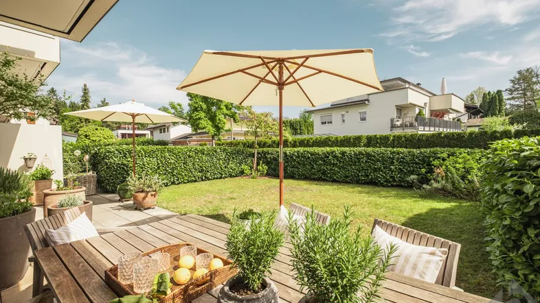 Große Terrasse mit Esstisch und Sonnenschirm, umgeben von einem gepflegten Rasen und Hecken.