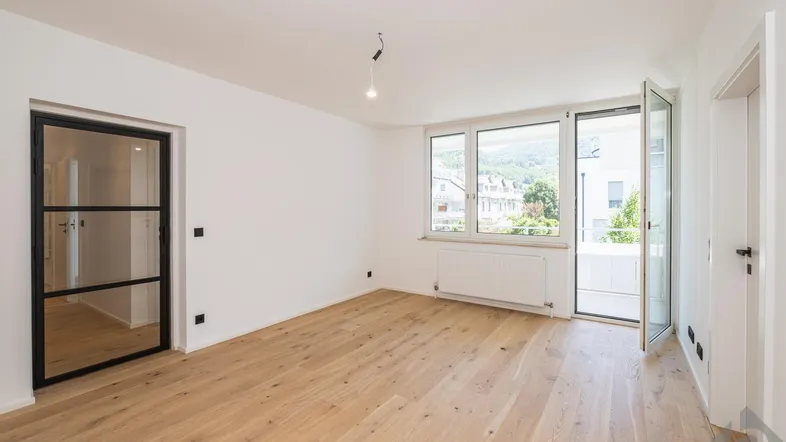 Helles Wohnzimmer mit Holzboden, Zugang zum Balkon und Blick auf die grüne Umgebung.
